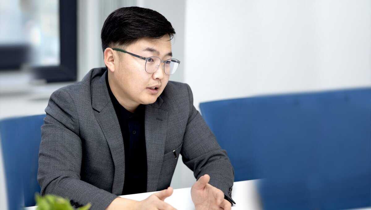 Саарал костюм, хар цамц өмссөн нүдний шилтэй хүн суугаад гараараа дохиж, бизнесийн яриа эсвэл хэлэлцүүлэгт оролцож байгаа мэт харагдана. Арын дэвсгэр дээр цэнхэр сандал, бүдэг оффисын орчин харагдаж байна. - Business.MN