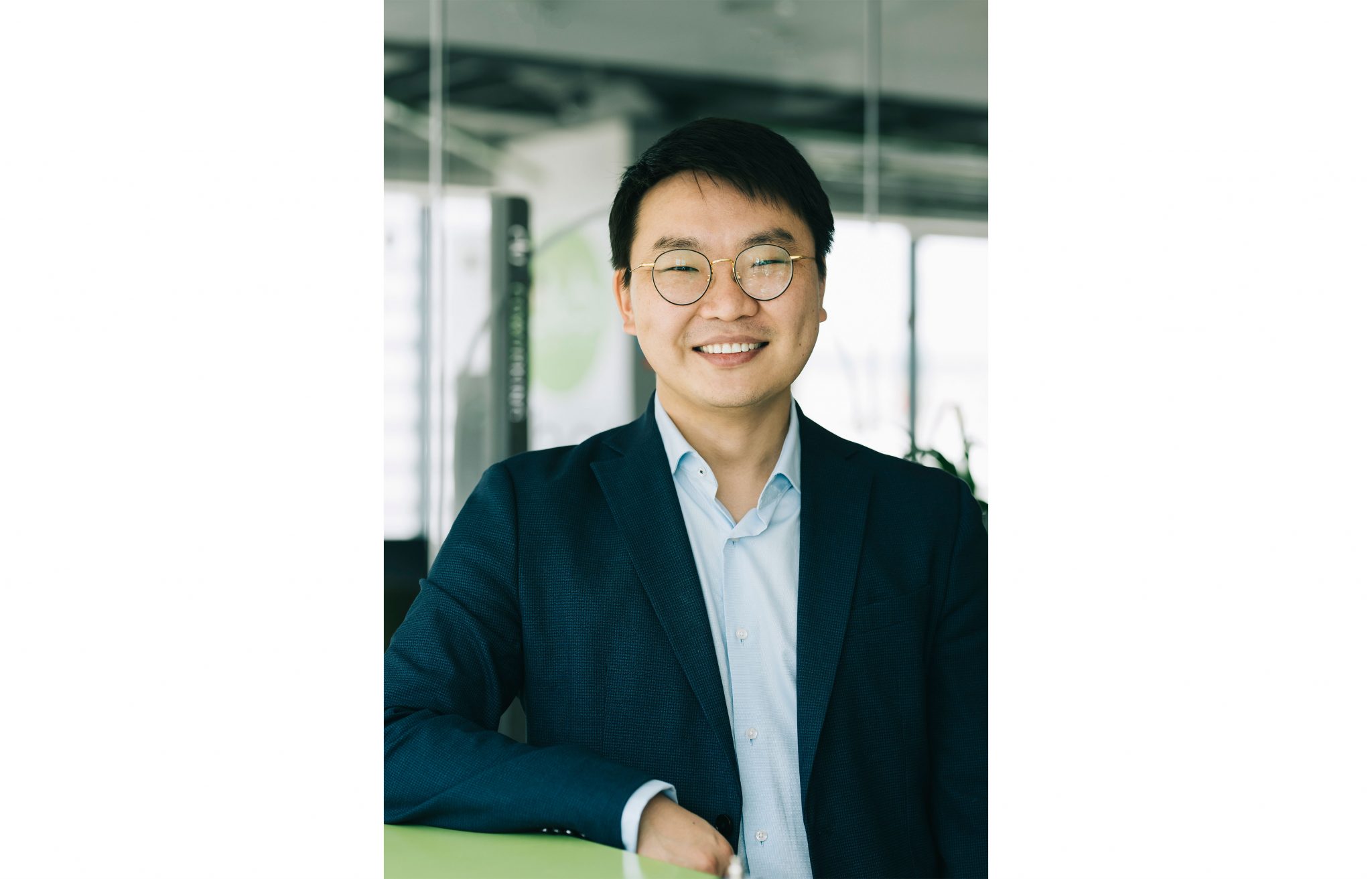 Нүдний шил, цэнхэр товчтой цамц, хар хөх өнгийн костюмтай хүрэм өмссөн хүн орчин үеийн офис орчинд зогсож байхдаа камер руу инээмсэглэж байна. Арын дэвсгэр дээр шилэн хана, бүдэг оффисын объектууд харагдаж байна. - Business.MN