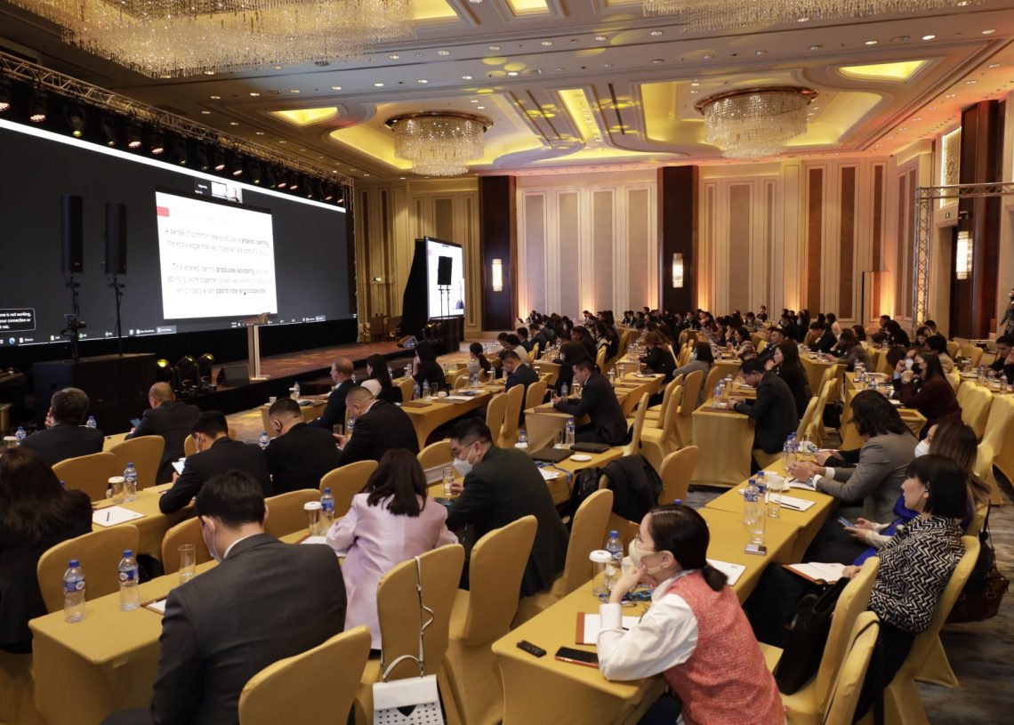 Шар ширээний бүтээлэгээр хучигдсан ширээний ард сууж буй бизнес эрхлэгчидээр дүүрэн хурлын танхим. Тэд өрөөний урд талд байрлах том дэлгэцэн дээр төвлөрч, үзүүлэнг үзүүлж байна. Тус байрыг гоёмсог лааны суурь, модон хавтангаар чимэглэсэн. - Business.MN