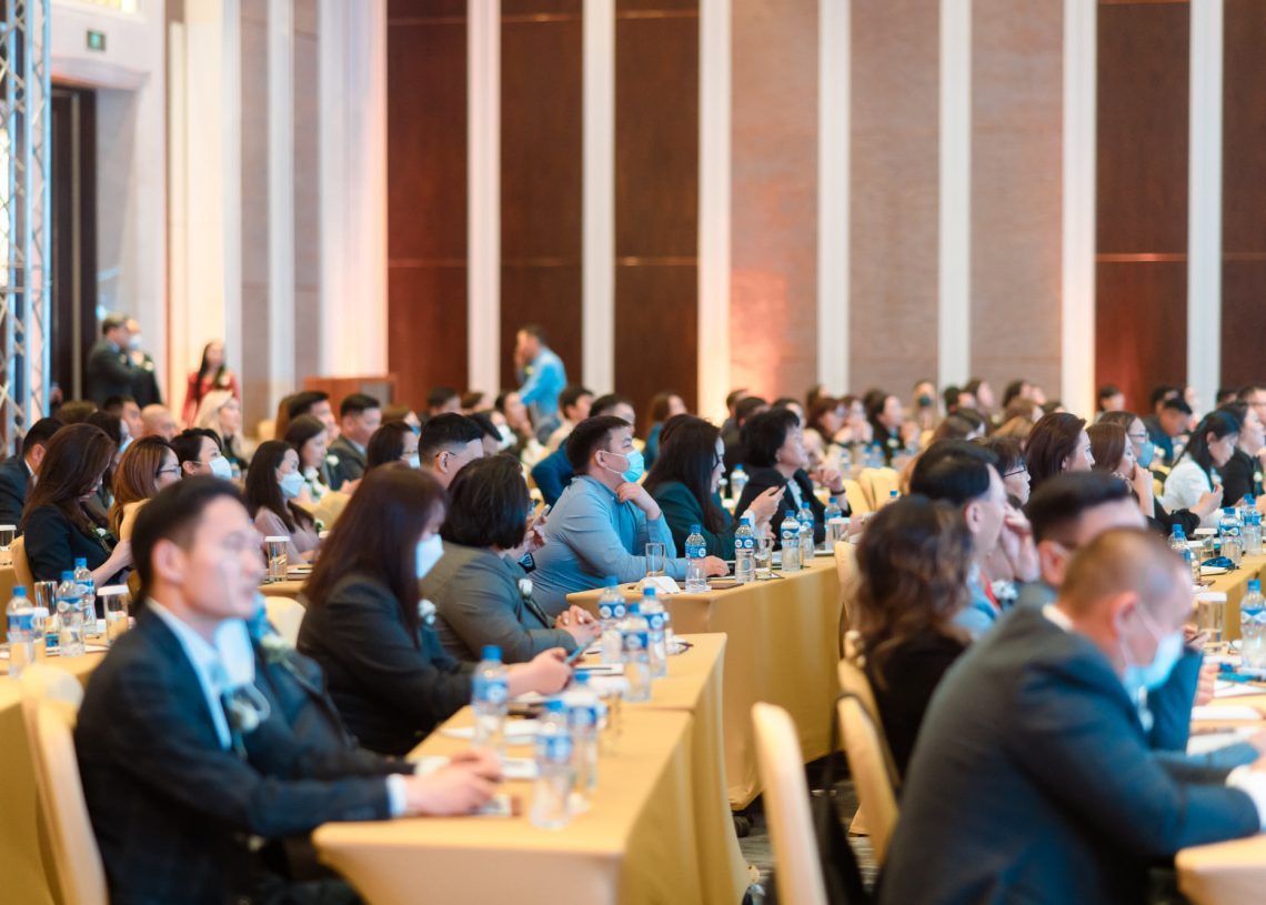 Хурлын танхимд олон тооны хүмүүс сууж, илтгэгчийн яриаг анхааралтай сонсож байна. Олон хүмүүс ажил хэргийн хувцас өмсөж, зарим нь маск зүүсэн байна. Өрөө нь шар бүрээстэй ширээ, савласан ус, тэмдэглэлийн дэвтэрээр дүүрэн байдаг. Энтрепренерүүд компанийн стратегийн талаар ярилцаж байх үед босоо хананы хавтангууд цаана нь харагдана. - Business.MN