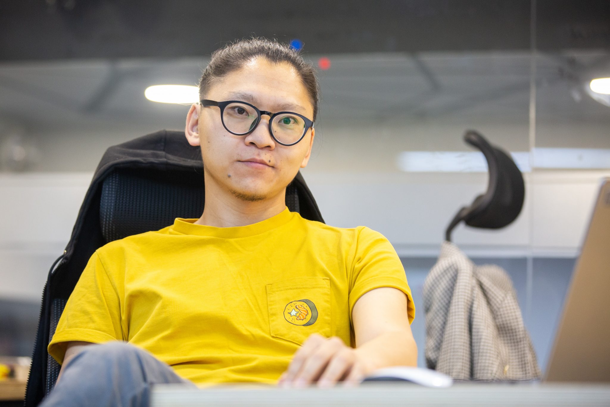 Нүдний шилтэй, үсээ боосон, жижиг дугуй лого бүхий шар цамц өмссөн бизнес-энтрепренер урд нь зөөврийн компьютертэй ширээний ард сууна. Цаана нь саарал хүрэм оффисын орчинд сандал дээр өлгөөтэй байна. - Business.MN