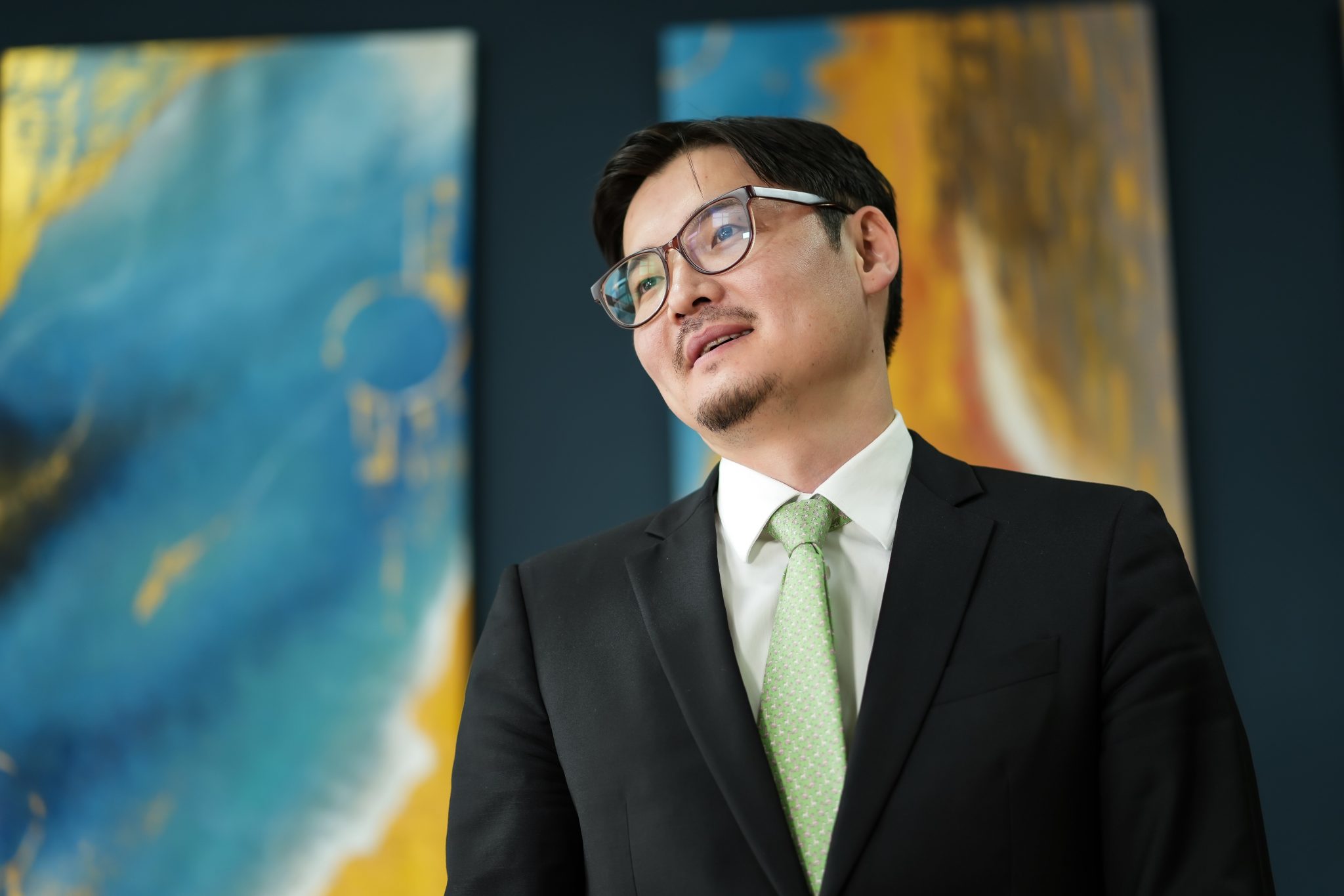 Хар костюм, цагаан цамц, ногоон зангиа өмссөн хүн хийсвэр өнгөлөг зургуудын өмнө зогсож байна. Тэр нүдний шил зүүж, толгойгоо үл ялиг хажуу тийш хазайлгаж, бизнесийн стратегийн талаар ярилцахад бэлэн энтрепренерийн өөртөө итгэлтэй зан чанарыг илтгэж, үл ялиг инээмсэглэж байна. - Business.MN