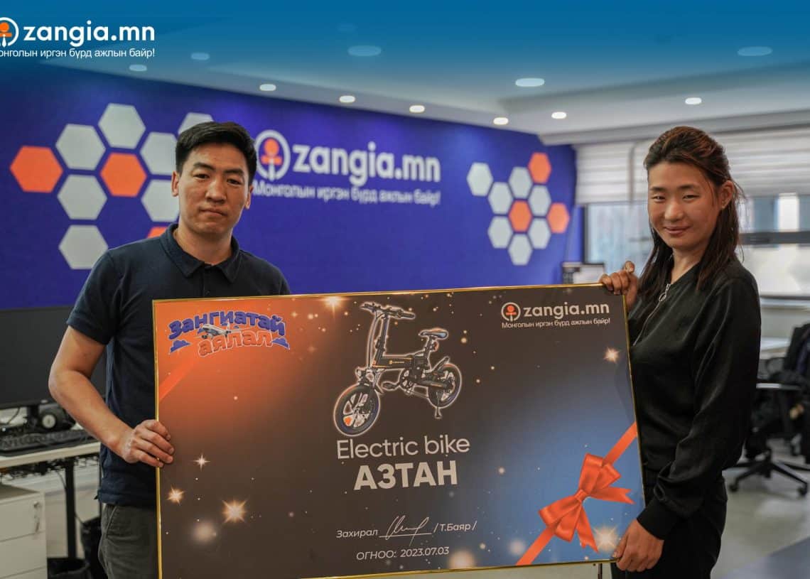 Хоёр хүн улаан туузаар чимэглэсэн том гэрчилгээ барьсан оффист зогсож байна. Энэхүү гэрчилгээнд цахилгаан дугуйны дүрс, англи хэл дээрх "Цахилгаан дугуй" гэсэн бичвэр, бусад монгол бичвэрийн хамт байрлуулсан байна. Тэдний ард ханан дээр zangia.mn сайтын баннер байрлуулсан нь компанийнхаа сүүлийн үеийн амжилтыг илэрхийлсэн байна. - Business.MN