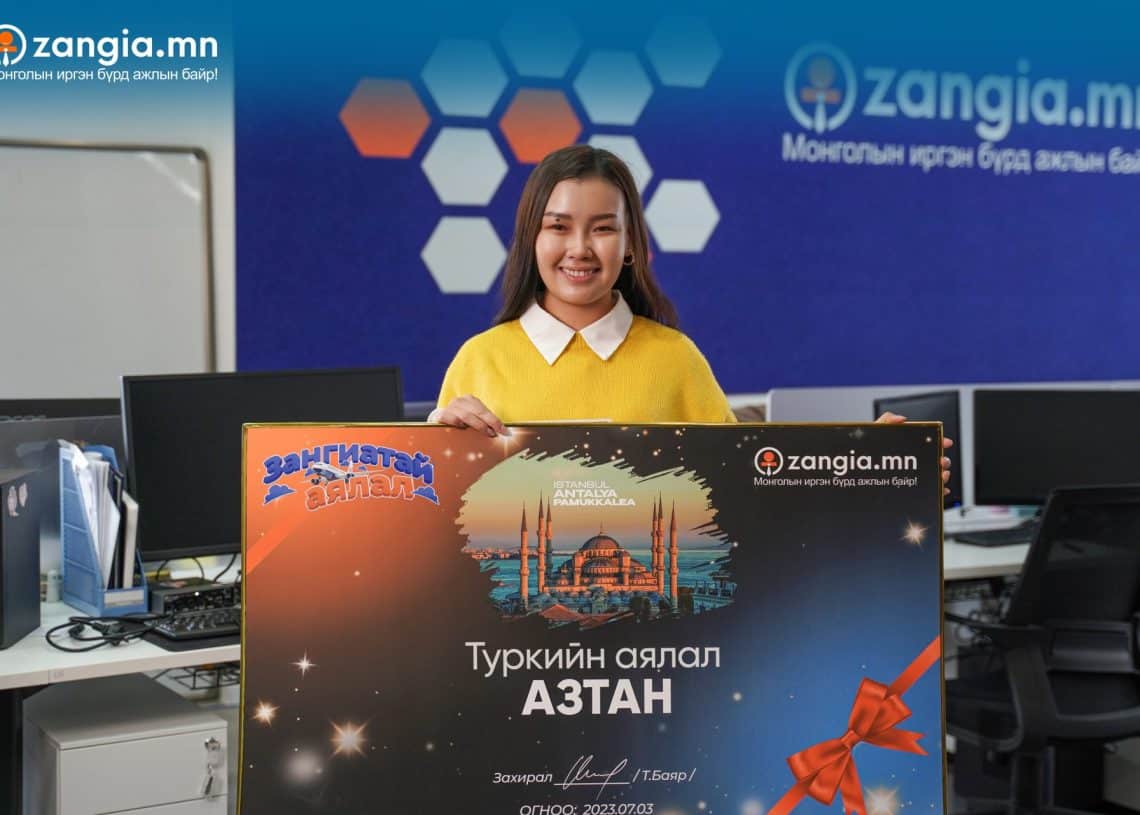 Монгол бичигтэй том чек барьсан шар ноосон цамцтай эмэгтэй оффисын байранд зогсож байна. Чек нь лого, тэнгэрийн шугам, бэлгийн туузны зургийг харуулдаг. Арын дэвсгэр дээр "Zangia.mn"-ийн лого харагдаж байна. Энэхүү энтрепренер нь компанийнхаа бизнесийн хүчин чармайлтын хүрээнд инновацийн сүнсийг харуулдаг. - Business.MN