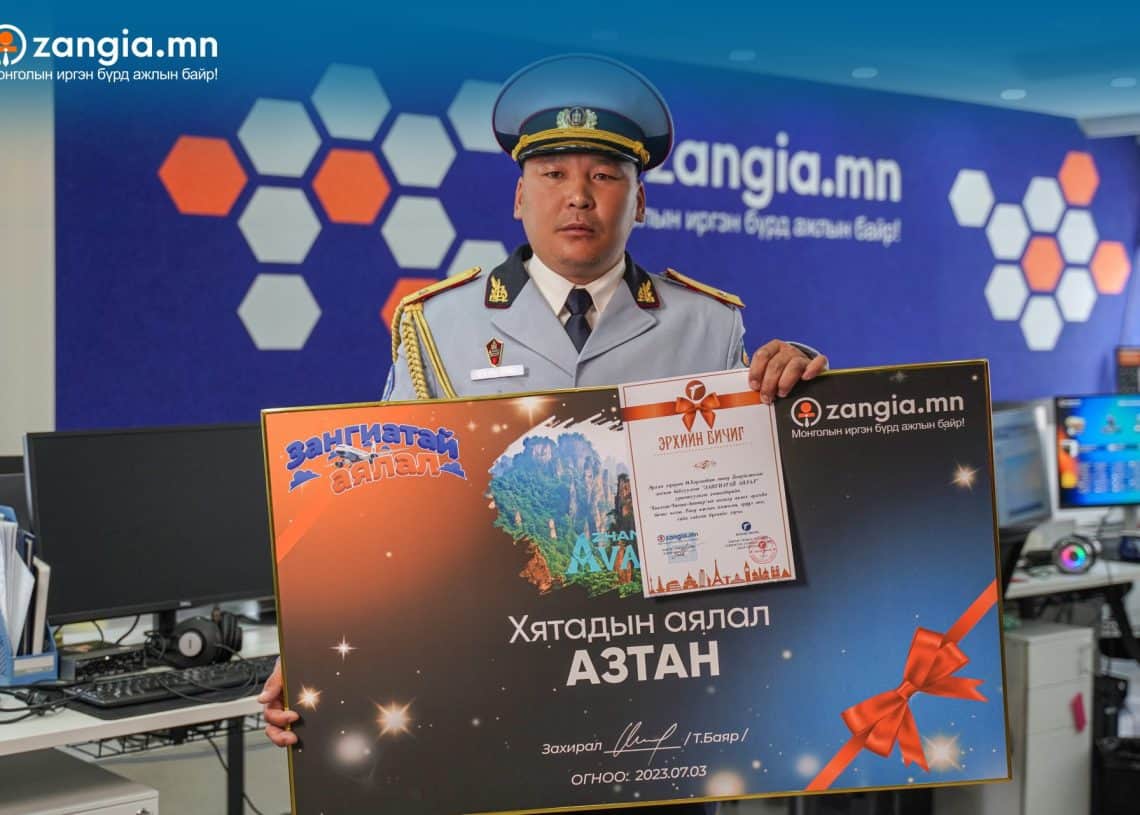 Дүрэмт хувцастай хүн монгол бичвэр бүхий том, өнгөлөг шагналын гэрчилгээ барьдаг. Тэрээр зургаан өнцөгт хээтэй, "zangia.mn", "zangia.com"-ын лого бүхий цэнхэр дэвсгэрийн өмнө зогсож байна. Арын дэвсгэр дээр бизнесийн ололт амжилтыг тэмдэглэсэн монгол бичгээр том бичвэртэй. - Business.MN