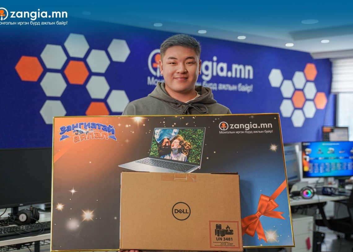 Оффис дээр зөөврийн компьютерын зураг, "zangia.mn" лого бүхий том бэлэг барьсан хүн зогсож байна. Баярын тууз, зүүн азийн бичгээр чимэглэсэн хайрцаг нь баяр ёслолын мөчийг сануулж байна. Ар талд нь хэд хэдэн компьютерийн дэлгэц харагдаж байгаа нь бизнесийн хөл хөдөлгөөн ихтэй орчныг илтгэнэ. - Business.MN