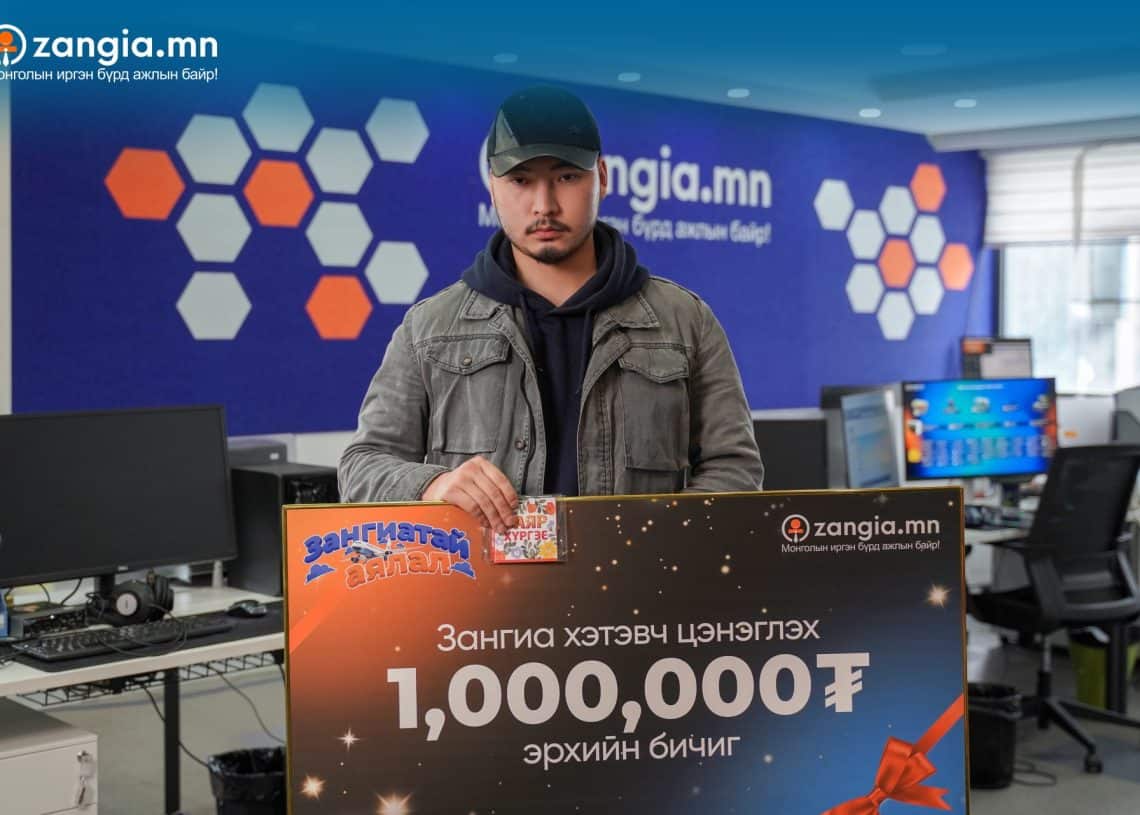 Зангиа.мн сайтаас 1,000,000₮-ийн их хэмжээний ёслолын чек бариад оффис дээр хар хүрэм, малгай өмссөн эрэгтэй зогсож байна. Арын дэвсгэр дээр компьютер бүхий ширээ, хананд нь Zangia.mn гэсэн брэндийг харуулсан нь компанийн бизнесийн идэвхтэй орчинг илтгэнэ. - Business.MN