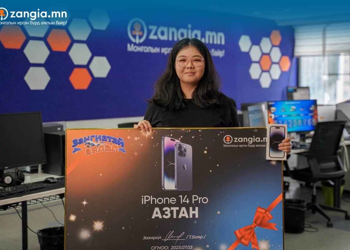 Урт хар үстэй, нүдний шилтэй эмэгтэй iPhone 14 Pro хожсон том сурталчилгааны тэмдэг барин оффист зогсож байна. Тэмдэглэгээний дээд талд Zangia.mn лого, iPhone-ийн зураг орсон байна. Цэнхэр өнгөөр чимэглэсэн оффис нь янз бүрийн ажилчдын ширээний ард сууж, бизнесийн динамик орчинг илэрхийлдэг. - Business.MN
