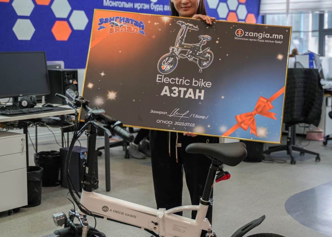 Инээмсэглэж буй бизнес эрхлэгч оффисын орчинд цахилгаан дугуй бэлэглэх том ваучер барьж байна. Ваучер нь дугуйн дүрс, компанийн лого болон бусад бичвэрийг агуулсан. Бизнес эрхлэгчийн өмнө гоёмсог цахилгаан дугуй байрлуулсан байна. - Business.MN