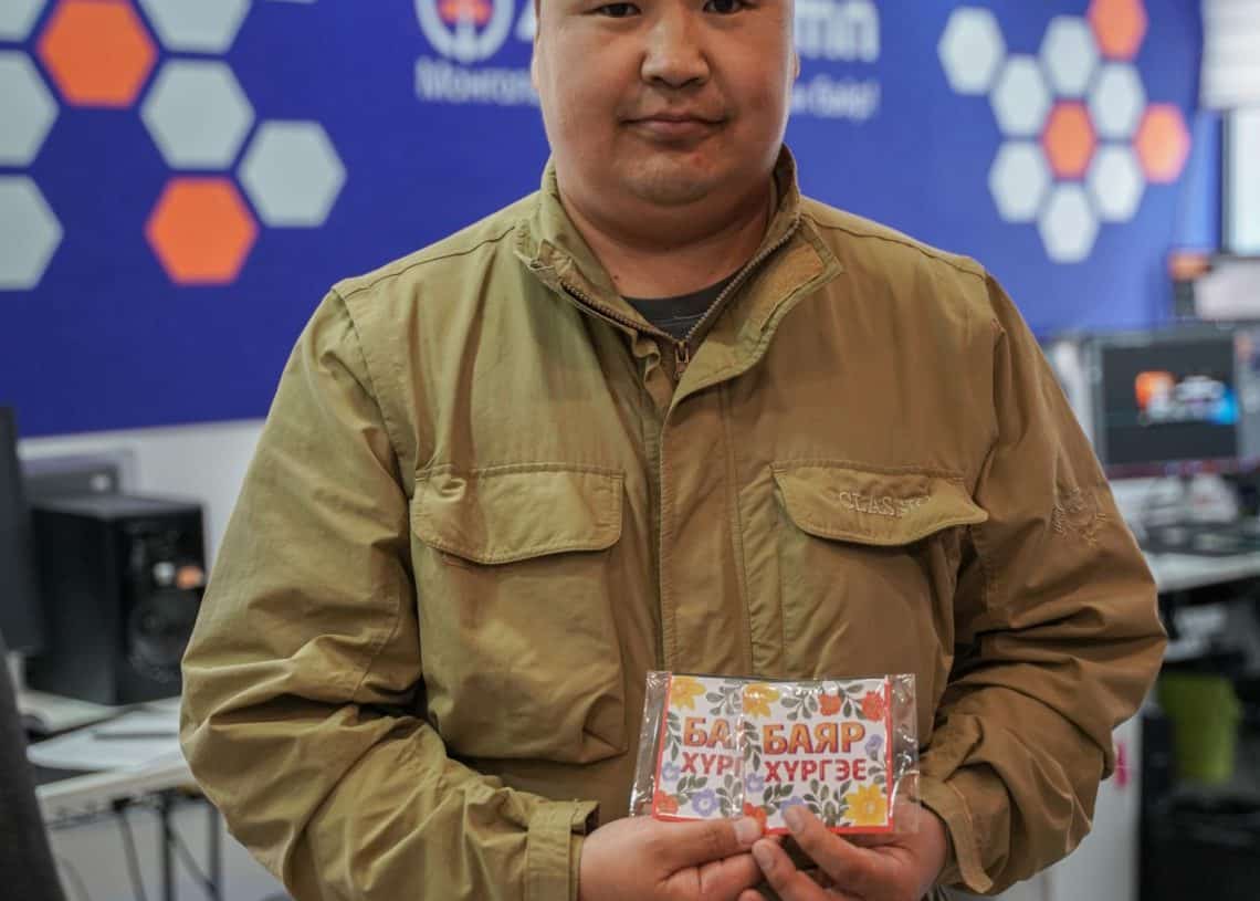 Нэг хүн оффисын өрөөнд хүрэн хүрэм өмсөж, хоёр боодол тоглоомын хөзөр барьдаг. Арын дэвсгэр дээр зургаан өнцөгт хээтэй цэнхэр, улбар шар өнгийн хана, компьютерийн тоног төхөөрөмж бүхий ширээ дүрслэгдсэн нь бизнес эрхлэгчийн бизнесийн ажлын байрны эрч хүчтэй эрч хүчийг илтгэнэ. - Business.MN