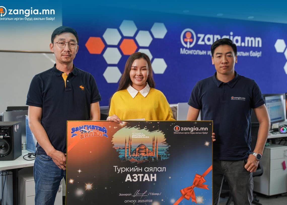 Гурван хүн оффисын дотор зогсож байна. Инээмсэглэн том ваучер барьсан голд байгаа хүн хажууд нь өөр хоёр хүн байна. Арын дэвсгэр дээр "zangia.mn" лого бүхий цэнхэр хана, компьютерийн ажлын байр зэрэг нь энэ эрч хүчтэй бизнест энтрепренерийн уур амьсгалыг цацруулж байна. - Business.MN