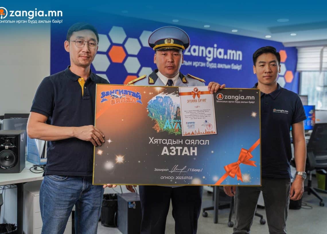 Гурван эр албан тасалгаанд "АЗТАН"-д хаягласан, баярын графикаар чимэглэсэн том шагналын үнэмлэхийг бардам барин зогсож байна. Нэг эрэгтэй хар цамцтай, нөгөө нь оройтой малгайтай дүрэмт хувцастай, гурав дахь нь хар өнгийн цамцтай. Тэдний ард бизнес эрхлэгчдийн ололт амжилтыг онцолсон "zangia.mn" баннер байдаг. - Business.MN