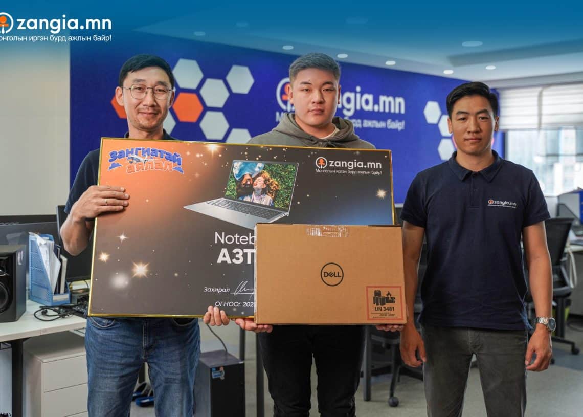 Гурван хүн дотор зогсож байна; голд байгаа хүн нь Dell зөөврийн компьютерын хайрцаг барьж байгаа бол зүүн талд нь "Zangia.mn Notebook" гэсэн том самбар харагдана. Арын дэвсгэр дээр Zangia.mn сайтын брэнд, оффисын тоног төхөөрөмж нь цэцэглэн хөгжиж буй бизнесийн орчныг харуулсан. - Business.MN
