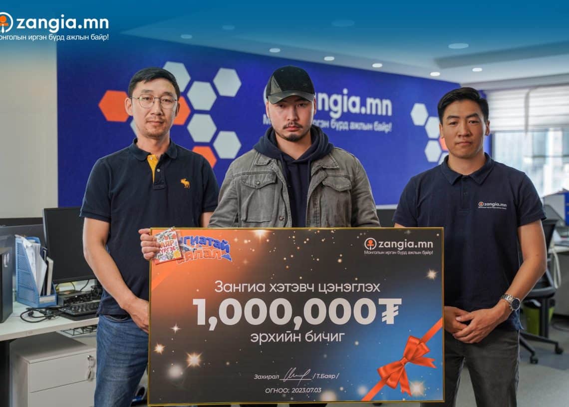 Гурван энтрепренер оффис дээр зогсож, тууз, нум бүхий ёслолын томоохон чек барьж байна. "zangia.mn" сайтаас авсан чек нь 1,000,000 Монгол төгрөгт зориулагдсан. Арын дэвсгэр дээр компанийн брэнд болон компьютерийг харуулсан. - Business.MN