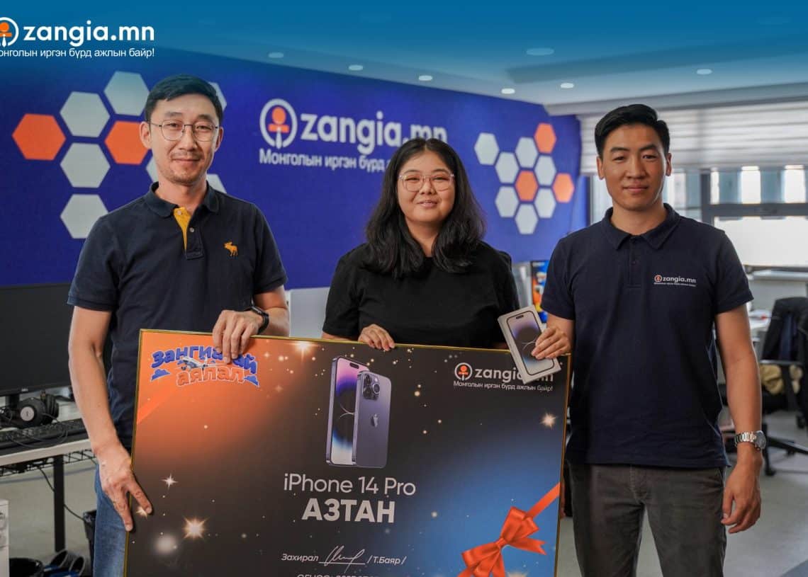 Гурван хүн "iPhone 14 Pro" гэсэн бичигтэй том зурагт хуудас барьж, шагналыг бэлгэддэг. Зурагт хуудас нь төхөөрөмжийн зургийг оруулсан болно. Арын дэвсгэр дээр цэнхэр хананд "zangia.mn" гэсэн хоёр лого бий. Инээмсэглэж, энгийн хувцасласан эдгээр энтрепренерүүд амжилтаа тэмдэглэж байна. - Business.MN