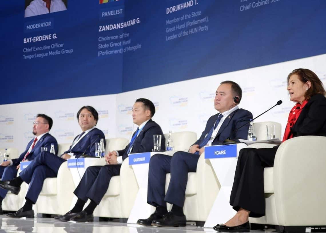 Таван хүн тайзан дээр сууж, хэлэлцүүлэгт оролцож байна. Тэдний дөрөв нь костюм, зангиа зүүсэн хоёр нь эрэгтэй, гурав нь анхааралтай сонсож, нэг нь чихэвчтэй. Улаан ороолт, бараан пиджак өмссөн нэг эмэгтэй микрофонд өөрийнхөө энтрепренерийн талаар ярьж байна. - Business.MN