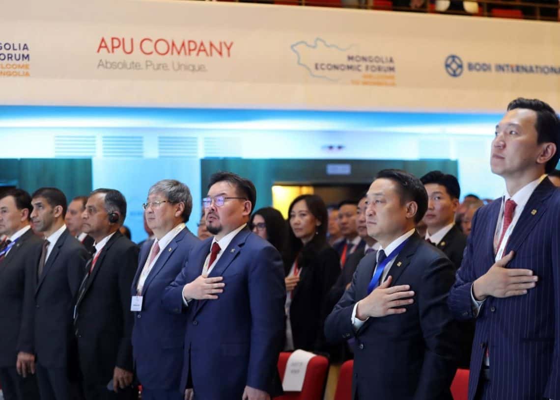 Хэсэг хүмүүс, тэр дундаа нэр хүндтэй энтрепренерүүд нэгэн арга хэмжээнд баруун гараа зүрхэн дээрээ бариад урагшаа харан зогсож байна. Тэд албан ёсны хувцас өмсөж, анхааралтай харагддаг. Цаана нь Монголын эдийн засгийн форумын сурталчилгааны баннерууд, янз бүрийн бизнесийн ивээн тэтгэгчдийг дээд талд нь байрлуулсан байна. - Business.MN