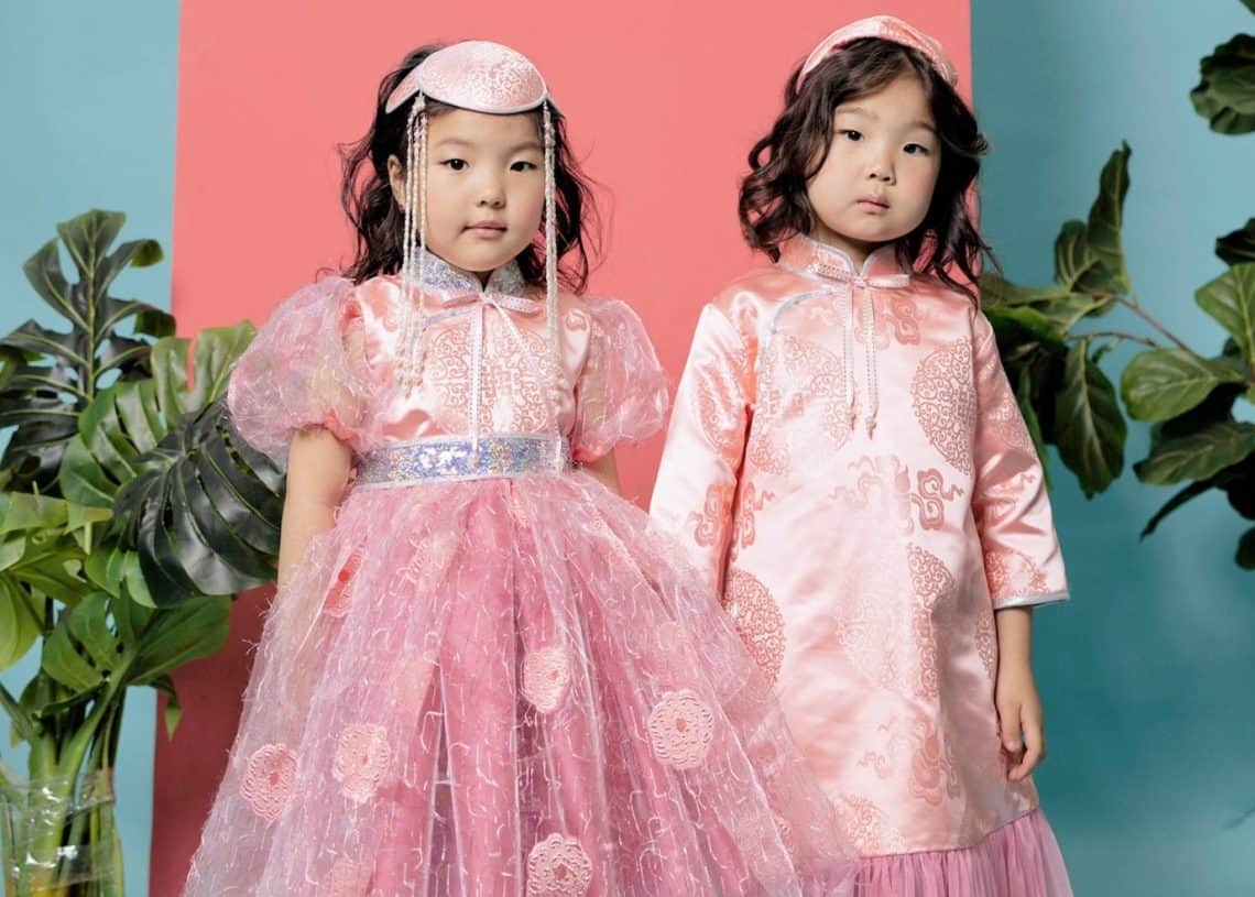 Ягаан нуман хаалга, цэнхэр дэвсгэрийн өмнө хоёр залуу охин зэрэгцэн зогсож байна. Тэд цэцгийн хээтэй, толгойндоо таарсан уламжлалт ягаан даашинз өмсдөг. Зүүн талд байгаа охин илүү хийсвэр даашинзтай бол баруун талд байгаа охин энтрепренер болохыг мөрөөддөг байж магадгүй тулай энгэртэй шулуун даашинзтай. - Business.MN