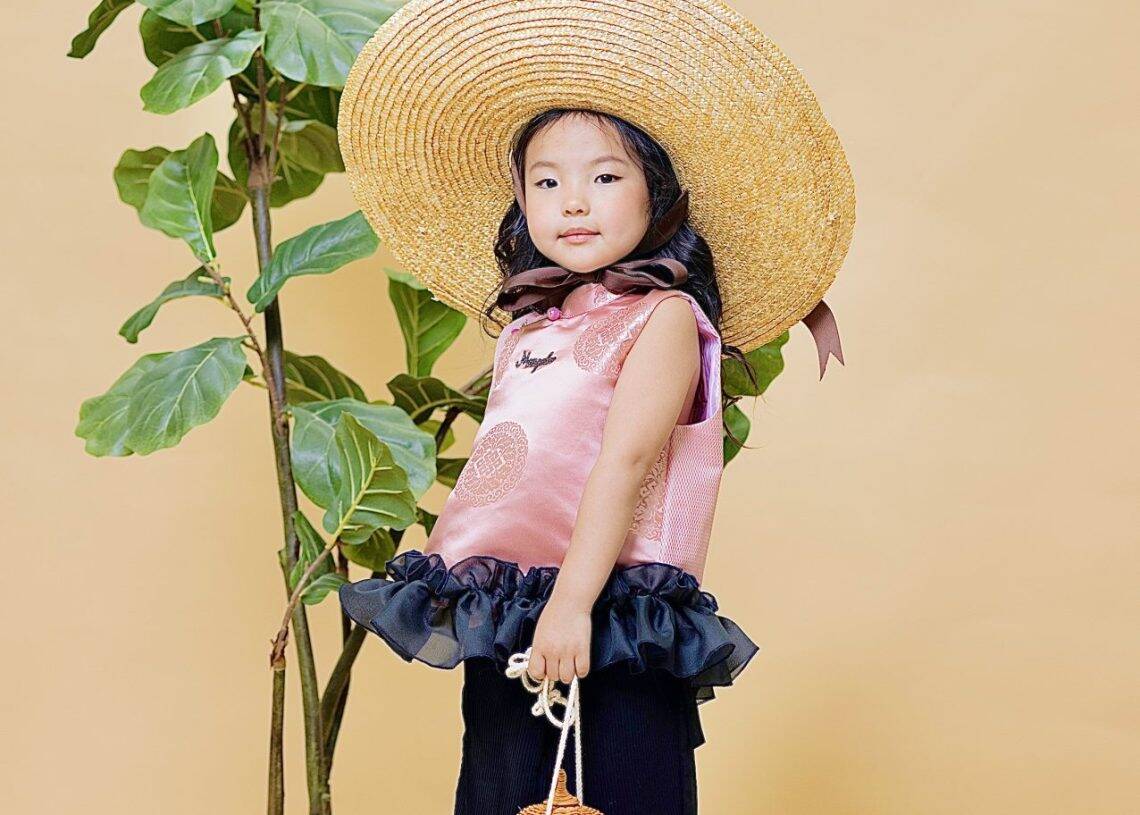 Залуу охин шаргал өнгийн дэвсгэр дээр зогсож байна. Том саравчтай малгай, ягаан өнгийн ханцуйгүй дээд тал нь дэрвэгэр нарийн ширхэгтэй, энгэртэй хар өмд, ягаан гутал өмсдөг. Тэр нэг гартаа жижиг сагс барьдаг бөгөөд энэ нь энтрепренерийн авъяас чадварыг харуулсан юм. Ойролцоох ваартай ургамал, зэгсэн хөөс. - Business.MN