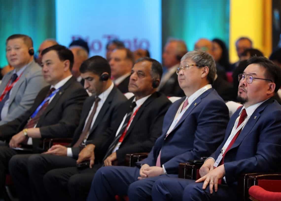 Хэсэг хүмүүс, голдуу албан ёсны ажил хэргийн хувцас өмссөн эрчүүд сандал дээр суун, анхааралтай сонсож байна. Тэд бизнесийн хурал эсвэл арга хэмжээнд оролцож байгаа бололтой, зарим нь орчуулга хийх гэж чихэвч зүүсэн байж магадгүй. Арын дэвсгэр нь цэнхэр, ногоон гэрэлтүүлгийн холимог юм. - Business.MN