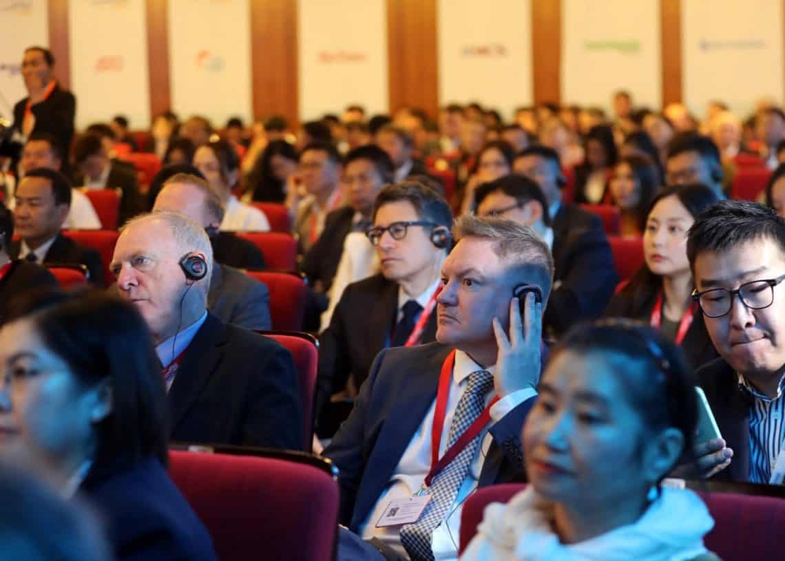 Хурлын өрөөнд олон янзын хүмүүс сууж байв. Олон оролцогчид, түүний дотор хэд хэдэн энтрепренерүүд чихэвч зүүж, анхааралтай сонсож байна. Өрөө нь улаан сандалаар дүүрсэн бөгөөд арын ханан дээр компанийн янз бүрийн лого харагдаж байна. Уур амьсгал нь албан ёсны бөгөөд төвлөрсөн байдаг. - Business.MN