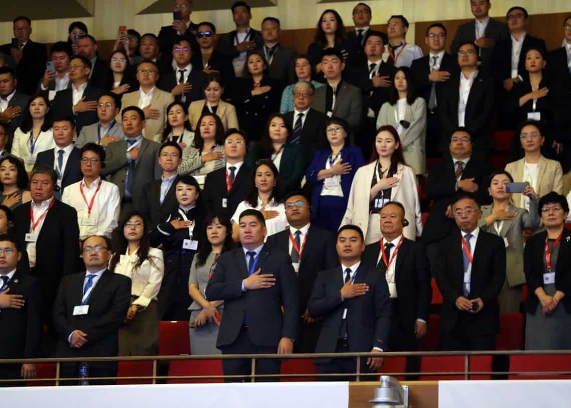 Албан ёсны хувцастай олон тооны хүмүүс танхимд зогсож байна. Олон хүмүүс баруун гараа зүрхэн дээрээ тавьж, эв нэгдэлтэй байх мөчийг харуулдаг. Спортын нэрийн шошго, эдгээр анхааралтай, хүндэтгэлтэй бизнесийн мэргэжилтнүүдийн аль аль нь entprerenerы компанийг багтаадаг, зорилго, зориулалтын мэдрэмжээр өрөө дүүргэх. - Business.MN