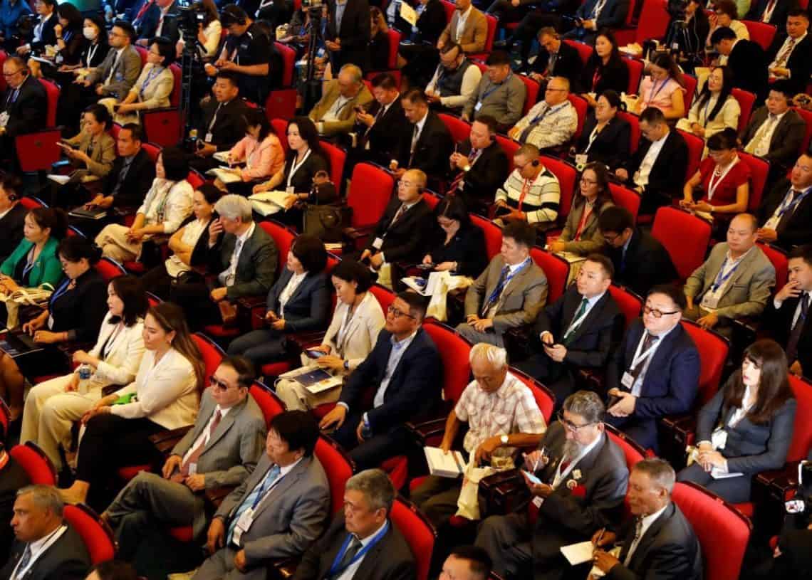 Албан ёсны хувцас өмссөн олон тооны хүмүүс улаан сандал дээр сууж, бизнесийн арга хэмжээ эсвэл чуулганд оролцож байна. Зарим нь бичиг баримт эсвэл цахим төхөөрөмж барьж байхад зарим нь харилцан яриа өрнүүлдэг. Үзэгчид анхааралтай, төвлөрсөн харагдаж байна. - Business.MN