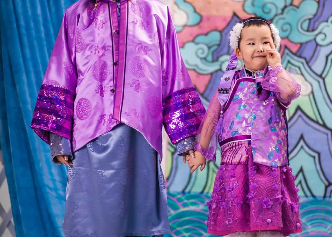 Нил ягаан, цэнхэр өнгийн уламжлалт, өнгөлөг хувцас өмссөн эмэгтэй, бага насны хүүхэд хоёр хөтлөлцөн зогсож байна. Хүүхэд жижиг цагаан тавцан дээр инээмсэглэн зогсож байна. Эргэдэг үүл дизайн бүхий эрч хүчтэй фон нь соёлын өвийг бизнес эрхлэх сэтгэлтэй хослуулан дүр төрхийг бүрдүүлдэг. - Business.MN