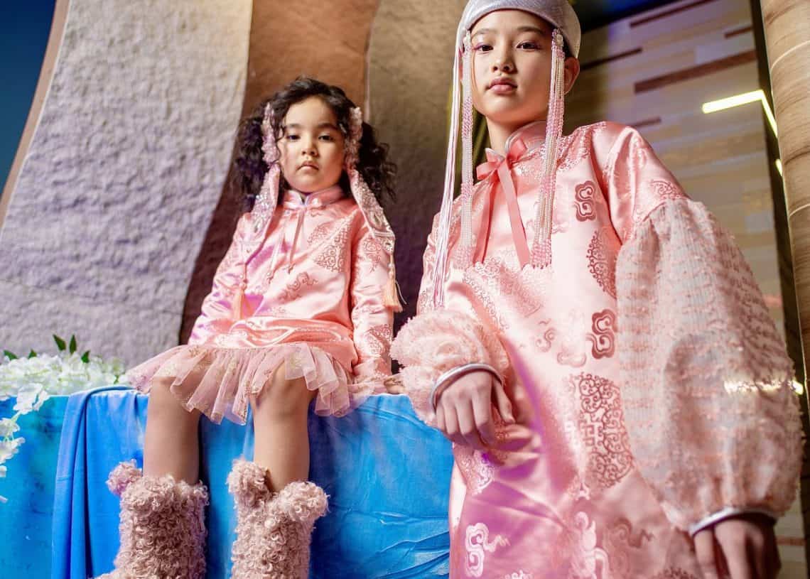 Чимэглэсэн ягаан өнгийн уламжлалт хувцастай хоёр хүүхэд дотор нь зургаа авахуулж байна. Зүүн талд байгаа хүүхэд цэнхэр даавуугаар хучигдсан гадаргуу дээр сууж, сэвсгэр ягаан гутал, цайвар ягаан, сэвсгэр даашинз өмсдөг. Баруун талд байгаа хүүхэд цагаан гоёл чимэглэлтэй урт ягаан даашинз өмсөж, соёлын арга хэмжээнд оролцож буй залуу бизнес эрхлэгчид шиг харагдаж байна. - Business.MN