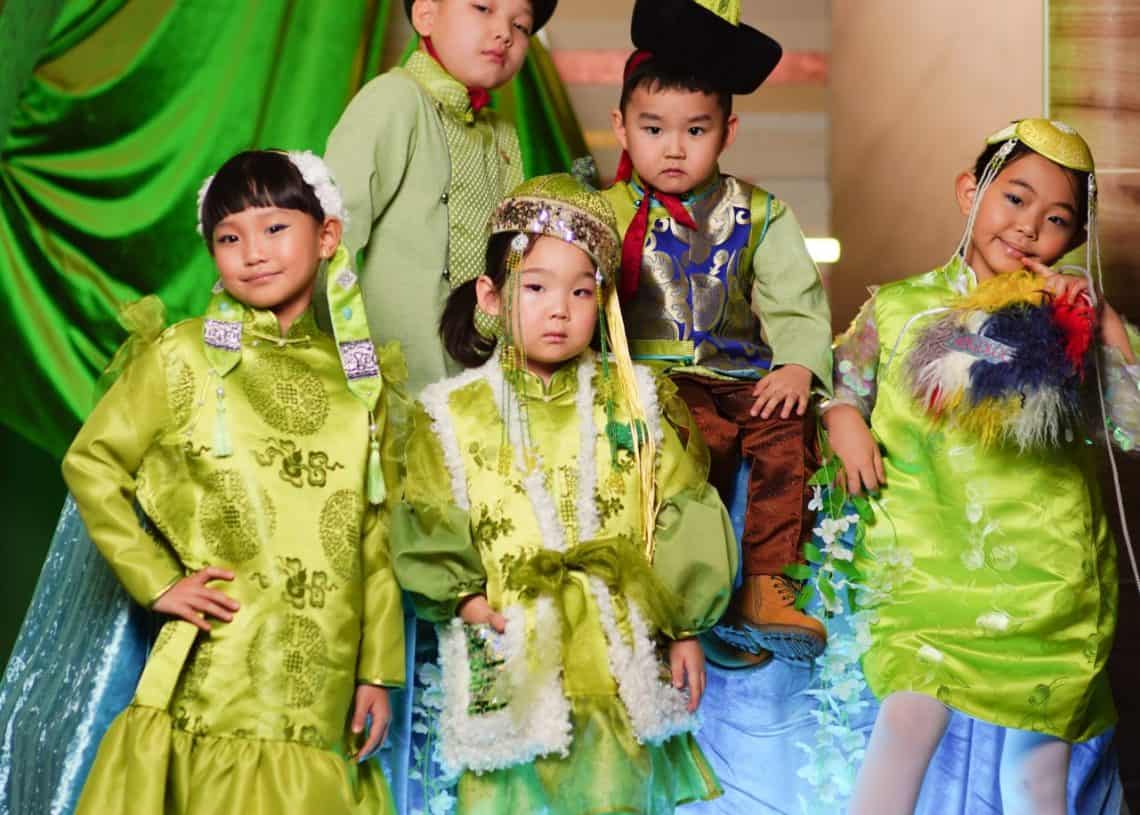 Уламжлалт хувцастай таван хүүхэд баярын уур амьсгалыг шингээсэн өнгөлөг фон дээр хөгжилтэй дүр үзүүлж байна. Тэд нарийн хээ, дагалдах хэрэгсэл бүхий тод ногоон өнгийн хувцас өмссөн байна. Арын дэвсгэр дээр ногоон хөшиг, цэнхэр даавуу багтсан нь баяр ёслолын үеэр залуу насны баяр баясгаланг нэмж өгдөг. - Business.MN