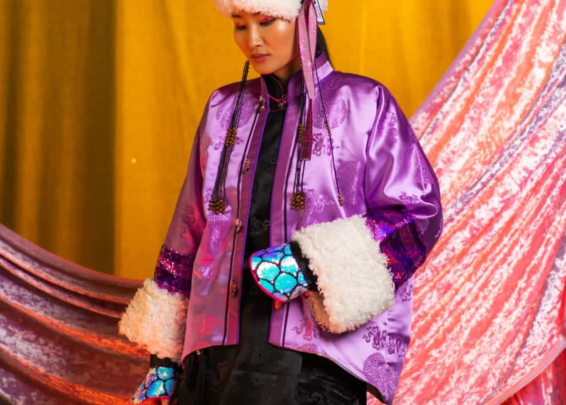 Нэг хүн монгол үндэсний хувцсаар гоёж, нарийн хийцтэй, цагаан үслэг ханцуйтай ягаан торгон хүрэм, гоёл чимэглэлийн малгай өмссөн байна. Арын дэвсгэрийг тод шар, ягаан өнгийн даавуугаар чимэглэсэн. Доод талд нь "Монголоо" компанийн лого харагдана. - Business.MN