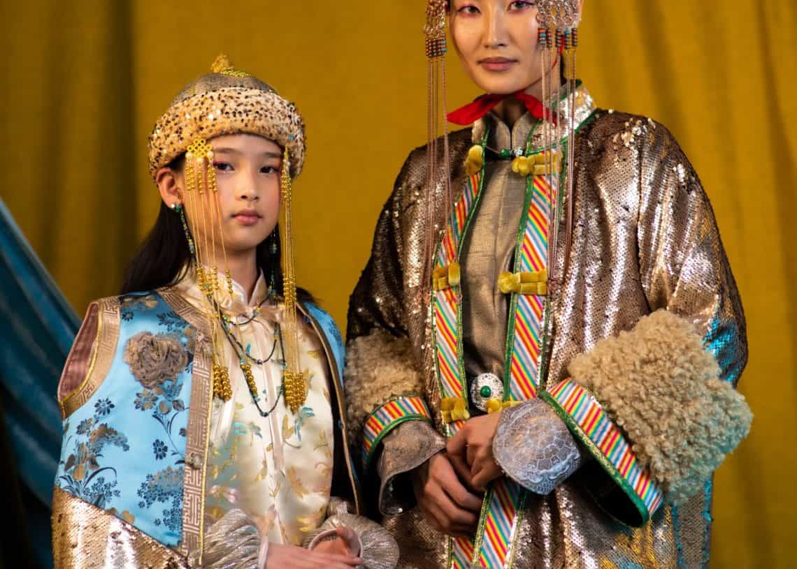 Алтан өнгийн дэвсгэрийн өмнө монгол үндэсний хувцас өмссөн хоёр хүн зогсож байна. Зүүн талд байгаа хүн үслэг дээл, малгай өмсдөг бол баруун талд байгаа хүн алтан толгойтой гялалзсан дээл өмсдөг. Хоёулаа нарийн ширхэгтэй дагалдах хэрэгсэл өмсдөг. Доод талд нь тэдний энтрепренер сэтгэлгээг харуулсан "Монголоо" усан тэмдэг бий. - Business.MN