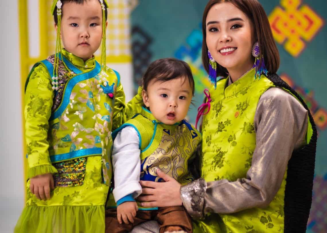 Цэнхэр хантааз, цагаан цамц өмссөн нялх хүүхдээ тэврэн хээтэй хивсэнцэр дээр тод ногоон уламжлалт хувцастай инээмсэглэсэн эмэгтэй сууж байна. Түүний хажууд ногоон даашинз өмссөн залуу охин зогсож байгаа бөгөөд энэ нь гэр бүлийн болон ирээдүйн бизнес эрхлэгчдийн урам зоригтой дүр төрхийг харуулсан хээтэй дэвсгэртэй өнгөлөг, уламжлалт өнгө аясыг нэмж байна. - Business.MN