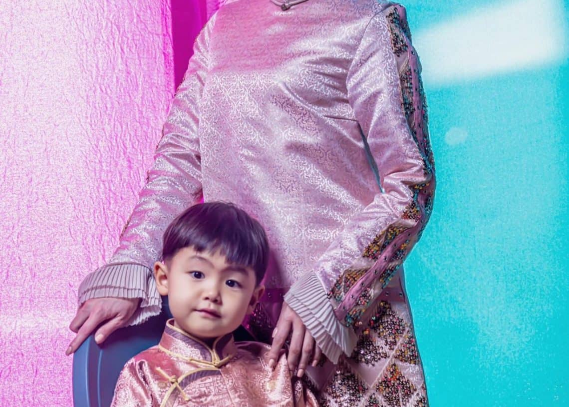 Сандал дээр сууж буй хүүхдийн ард уламжлалт ягаан хувцас өмссөн энтрепренер залуу эмэгтэй зогсож байна. Мөн хүүхэд цэнхэр өмдтэй ягаан уламжлалт хувцас өмссөн байна. Тэд хоёулаа инээмсэглэж, ягаан, оюу өнгө бүхий эрч хүчтэй дэвсгэр дээр байрлуулсан.
- Business.MN