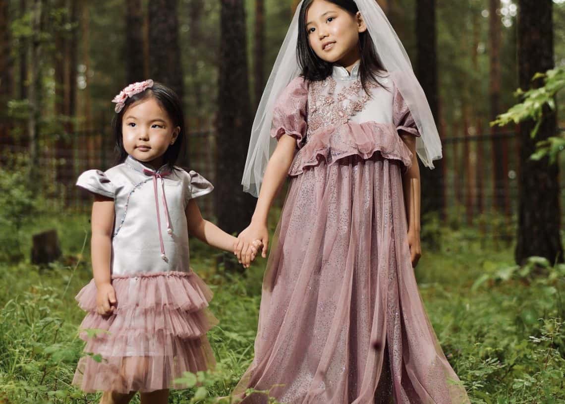 Хоёр залуу охин ойд хөтлөлцөн зогсож байна. Хөшигтэй цайвар ягаан даашинз өмссөн хөгшин охин энтрепренер болохыг мөрөөддөг байж магадгүй гэж бодсоор хажуу тийшээ харна. Ягаан өнгийн ороомогтой мөнгөн даашинзтай бага охин урагшаа харна. Тэдний эргэн тойронд өндөр мод, ногоон навчис хүрээлэгдсэн байдаг. - Business.MN