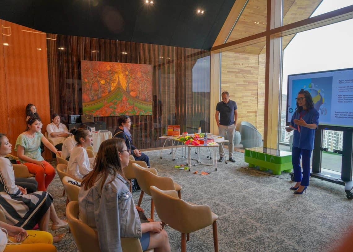 Хэсэг хүмүүс орчин үеийн шилэн ханатай өрөөнд суугаад том дэлгэцийн өмнө илтгэл тавьж буй эмэгтэйг анхааралтай сонсож байна. Өрөөнд модон өргөлт, өнгөлөг зураг, ширээн дээр тараагдсан гар урлалын хэрэгслүүд байдаг. Бизнесийн инновацийг удирдан чиглүүлж буй бизнес эрхлэгчийн сүнсийг агуулсан эр дэлгэцийн дэргэд зогсож байна. - Business.MN