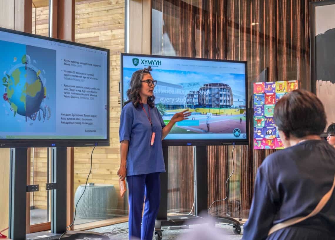 Боловсролын агуулгыг харуулсан хоёр том дэлгэцийн өмнө нэгэн эмэгтэй зогсоод ярьж байна, нэг нь дэлхийн график, нөгөө нь барилгын зураг. Хүмүүс тэргүүн эгнээнд сууж, энтрепренер болох хүсэлтэй хүмүүст зориулсан бизнесийн стратегийн талаарх түүний ойлголтыг сонсож байна. - Business.MN