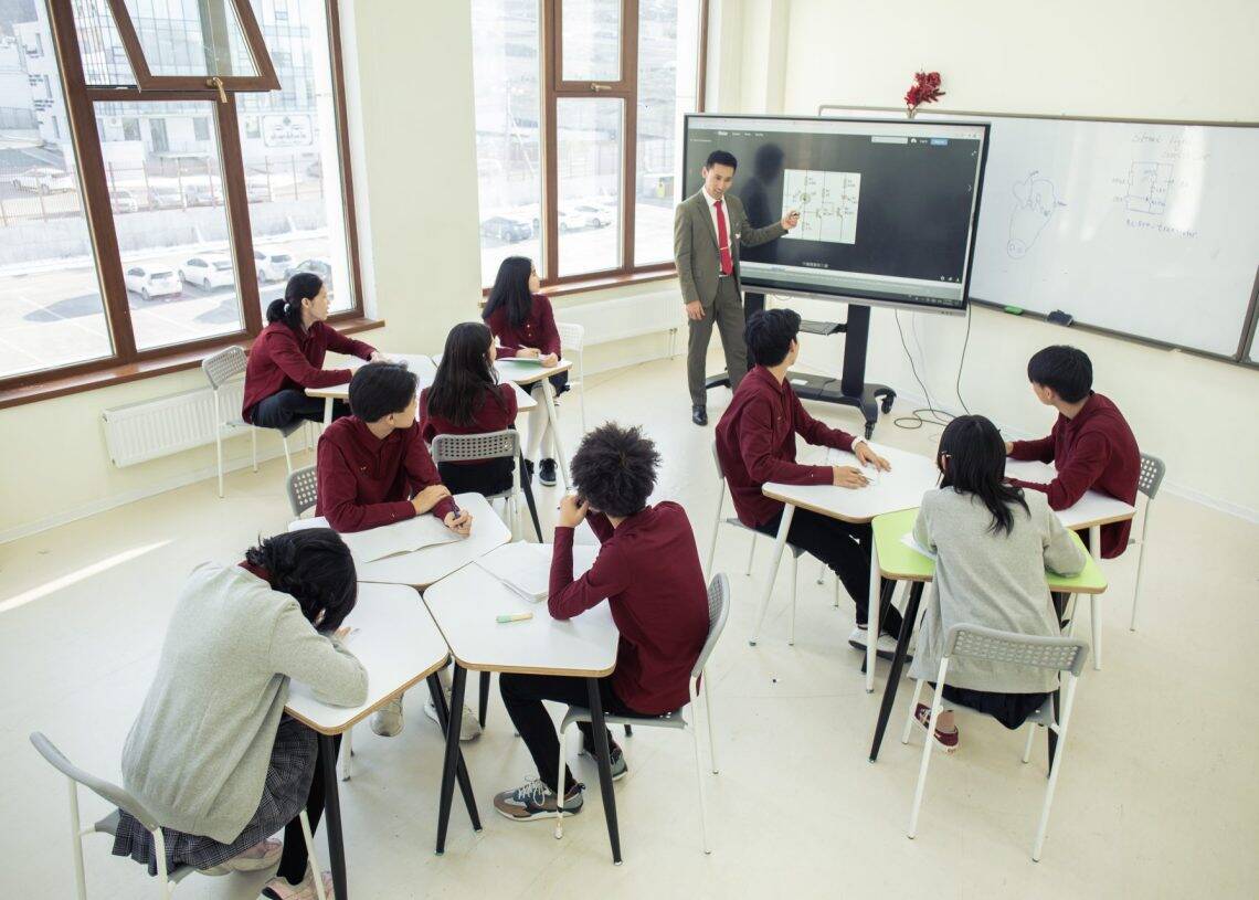 Хагас тойрог хэлбэрээр ширээн дээр суугаад том дэлгэцээр илтгэл тавьж буй багшийг анхааралтай ажиглаж буй оюутнуудтай анги. Багш дэлгэц рүү зааж, бизнесийн зарчмуудыг ярилцаж, хүрэн улаан өнгийн дүрэмт хувцас өмссөн сурагчид тэмдэглэл хөтөлнө. Том цонх, самбар нь өрөөг чимдэг. - Business.MN