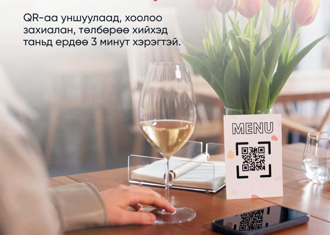 QR цэсийн картыг рестораны ширээн дээр цагаан дарс, ухаалаг утас, ягаан алтанзул цэцэг бүхий ваарны дэргэд байрлуулсан байна. Уг бичвэр нь монгол хэл дээр байгаа бөгөөд "aMENU" компани ухаалаг QR цэс ашиглан хоол захиалж, 3 минутын дотор төлбөрөө төлөхийг сурталчилж байна. "aMENU" лого дээд талд харагдаж байна. - Business.MN