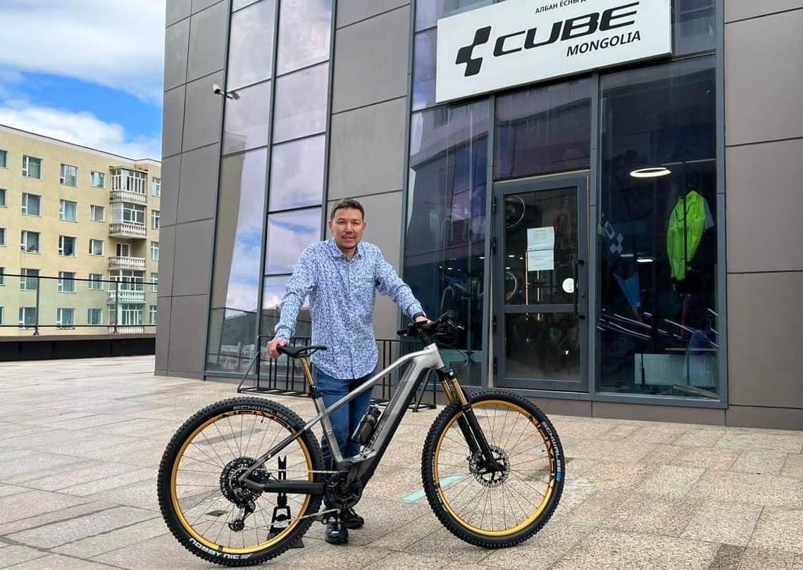 Хээтэй цамцтай хүн "Cube Mongolia" гэсэн бичигтэй барилгын урд хоббигоо тусгаж дугуйны хажууд зогсоно. Арын дэвсгэр нь үүлэрхэг тэнгэрийн доорх бусад барилгуудыг харуулж, хотын амьдралын мөн чанарыг харуулсан. - Business.MN