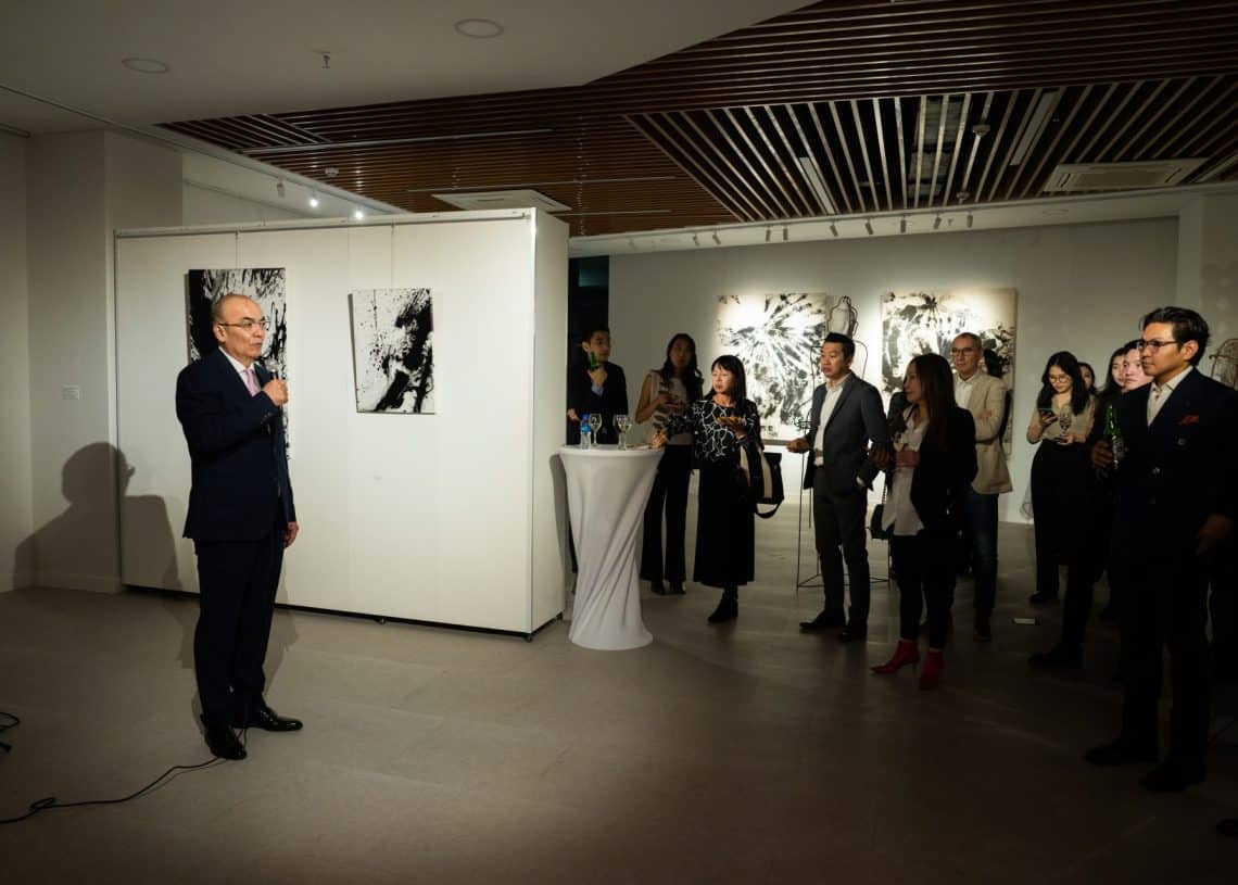 Костюм өмссөн хүн, магадгүй санхүүч, урлагийн галерейн арга хэмжээнд микрофоноор ярьж байна. Уран бүтээлээр чимэглэгдсэн өрөөнд хэд хэдэн хүн анхааралтай сонсож зогсоно. Ойролцоох ундаа бүхий жижиг ширээ байдаг бол таазны гэрэл нь эрч хүчтэй орон зайг гэрэлтүүлдэг. - Business.MN