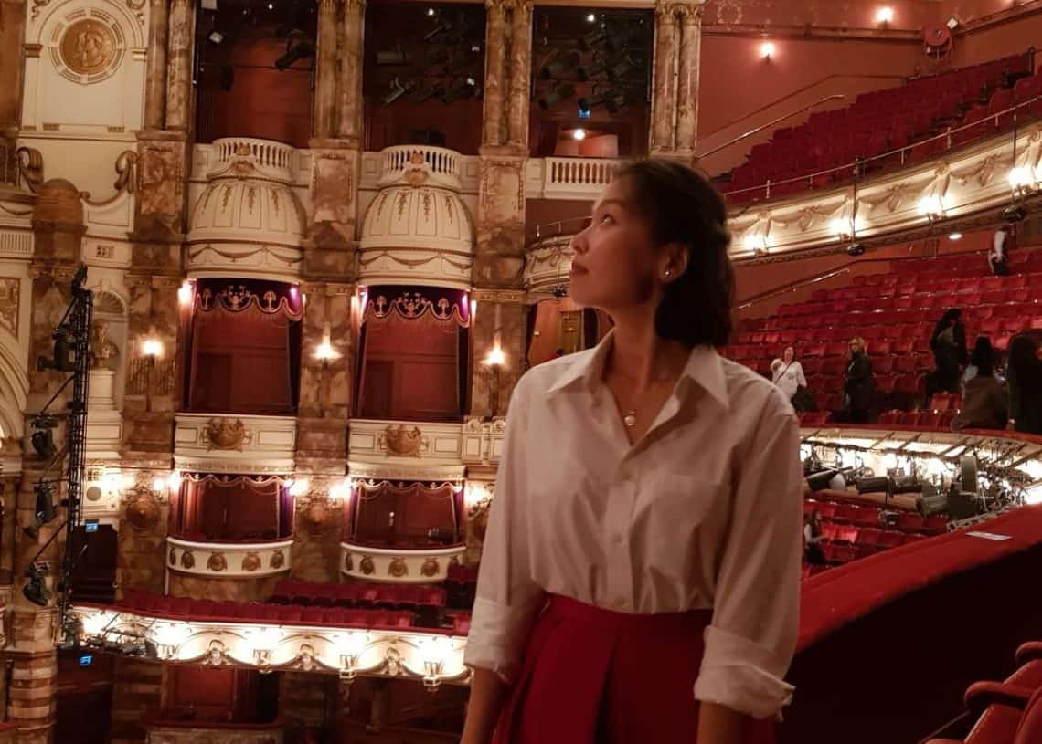 Цагаан цамц, улаан юбка өмссөн, урлагт элэгтэй, гоёмсог, хоосон театрын тагтан дээр зогсож буй хүн. Тэд улаан суудал, гоёл чимэглэлийн архитектураар хүрээлэгдсэн тайзны талбай руу ширтэж, урлаг сонирхогчдод ч, санхүүчдэд ч хамаатай гоо үзэсгэлэнд төөрсөн байна. - Business.MN