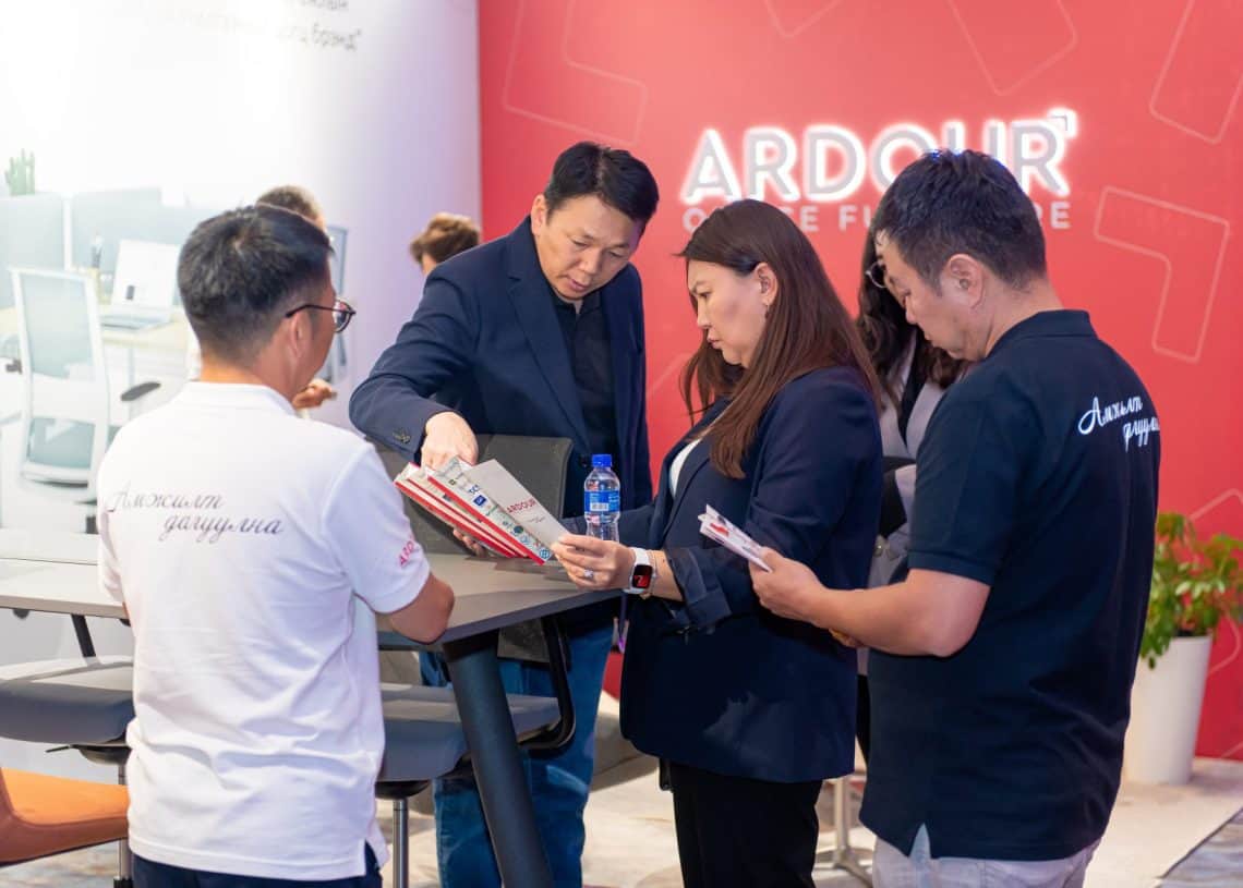 Дөрвөн хүний бүрэлдэхүүнтэй хэсэг хүмүүс ширээний ард цугларч, 2024 онд болох Монголын хөдөлмөрийн зах зээлийн дээд хэмжээний уулзалтын талаарх товхимолуудыг хэлэлцэж байна. Арын дэвсгэр дээр "ARDOUR OFFICE FURNITURE" лого бүхий улаан хана харагдана. Тэд анхаарлаа төвлөрүүлж, яриа өрнүүлж байгаа нь хүний нөөцийг сонирхож байгаагаа онцолж байна. - Business.MN