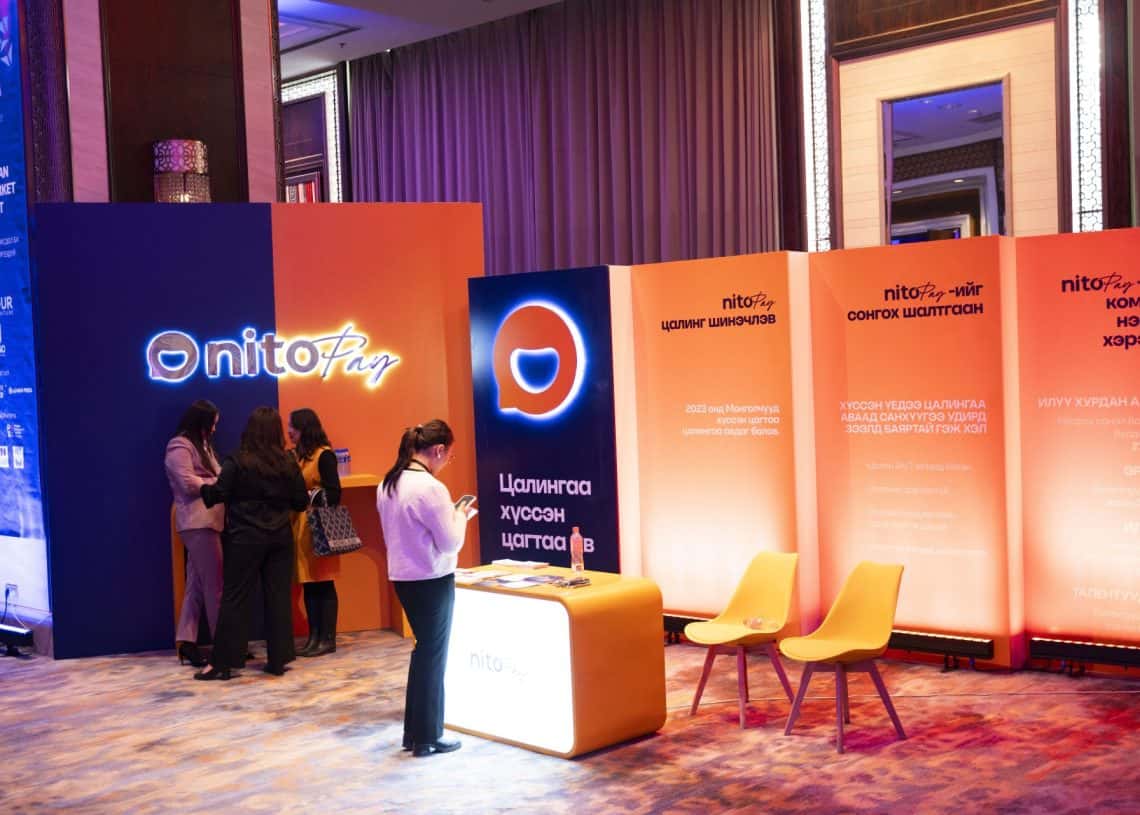 Хана, ширээн дээр "nito ань" гэсэн лого бүхий тод өнгийн үзэсгэлэнгийн лангуун дээр хэсэг хүмүүс харилцаж байна. Монголын хөдөлмөрийн зах зээлийн дээд хэмжээний уулзалт 2024 арга хэмжээний нэг хэсэг болох уг байгууламж нь орчин үеийн дотоод орчинд гэрэлтдэг самбар, суудлын талбай, мэдээллийн дэлгэц зэргийг багтаасан болно. - Business.MN