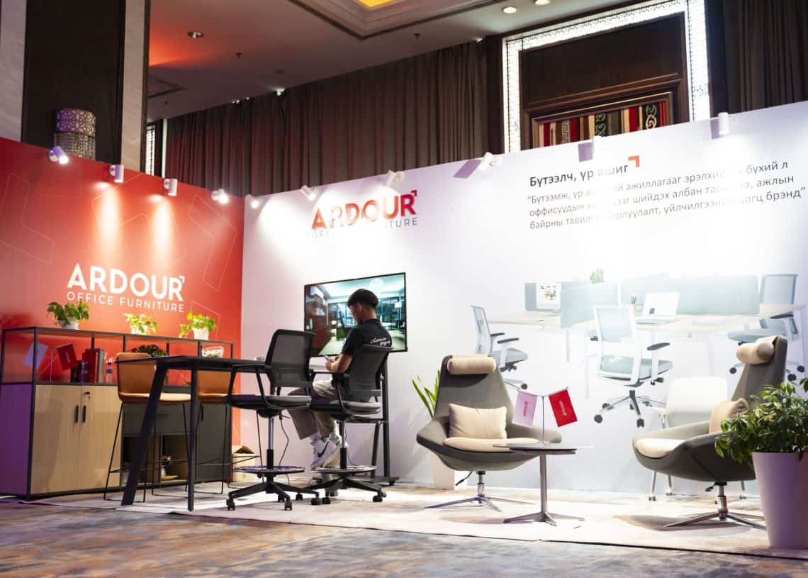 Монголын хөдөлмөрийн зах зээлийн дээд хэмжээний уулзалт 2024 дээр Эргономик оффисын сандал, ширээг харуулсан Ardor Office Furniture-ийн үзэсгэлэнгийн лангууг сонирхоорой. Мэргэжилтэн компьютертэй ширээний ард сууж, ханан дээр гоёмсог гэрэлтүүлэг, сурталчилгааны материалууд дунд сууж байна. - Business.MN