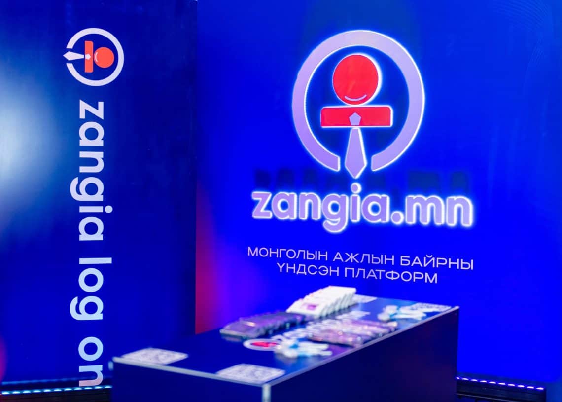 Цэнхэр гэрэлтүүлэгтэй өрөөнд "zangia.mn" лого нь монгол бичгээр бичсэн тэмдгийн хажууд тод гэрэлтдэг. Хүснэгтэнд хүний нөөцийн шийдлийг эрэлхийлж буй Монголын хөдөлмөрийн зах зээлийн дээд хэмжээний уулзалт 2024-т зориулсан сурталчилгааны материалыг харуулсан байна. - Business.MN