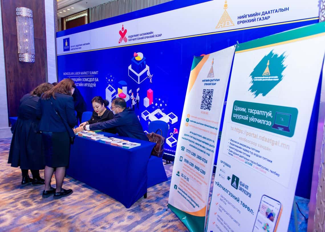 2024 оны дээд хэмжээний уулзалтын үеэр хэд хэдэн хүмүүс товхимол, мэдээллийн баннераар чимэглэсэн ширээний дэргэд цугларав. Баннерууд нь дижиталчлал, технологийн санаачлага, хөгжиж буй Монголын хөдөлмөрийн зах зээлтэй холбоотой бичвэр, логог онцолсон байна. Арын дэвсгэр дээр нэмэлт дэлгэрэнгүй мэдээлэл бүхий график дүрслэгдсэн байна. - Business.MN