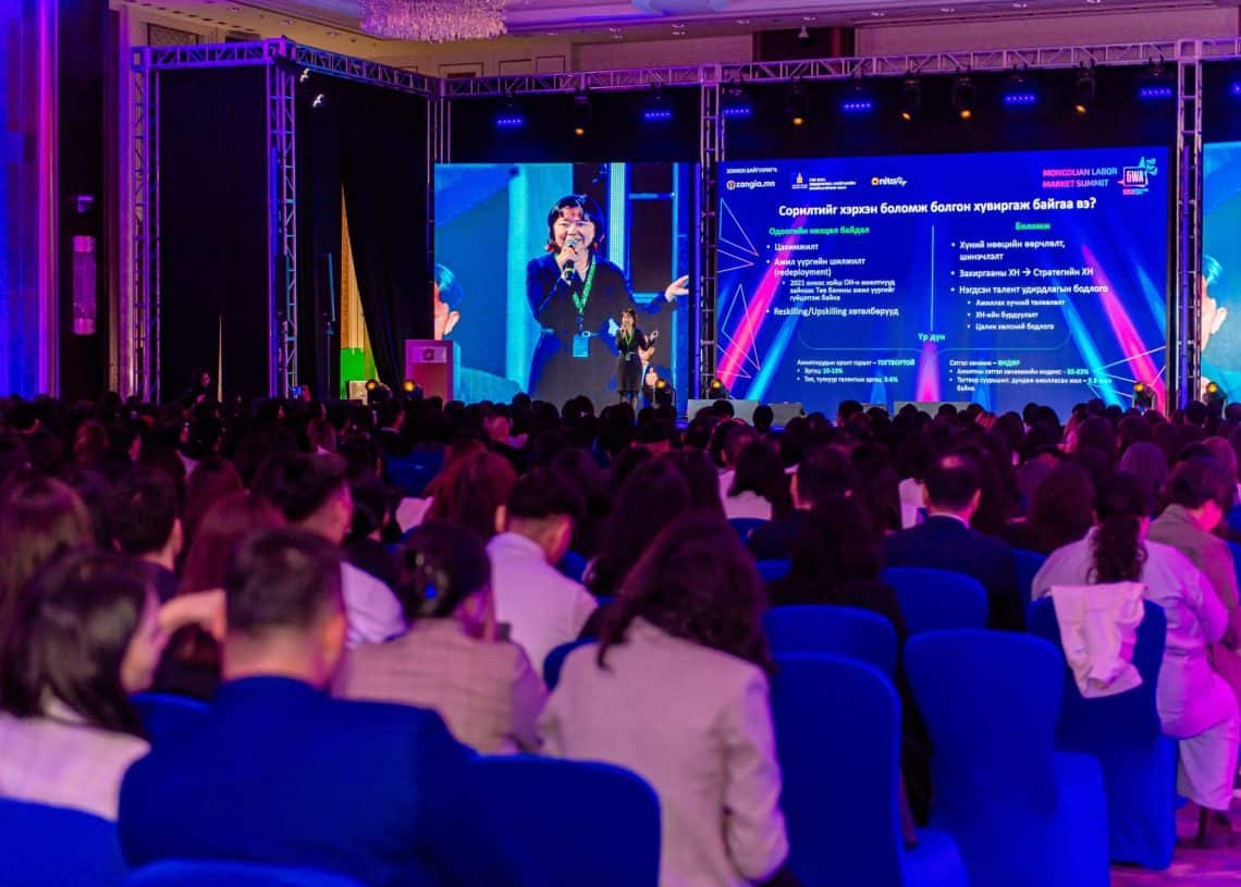 “Монголын хөдөлмөрийн зах зээлийн дээд хэмжээний уулзалт 2024” хурлын танхимд илтгэгч тайзан дээр зогсож, олон нийтэд хандаж байна. Өрөөний цэнхэр сандал нь дэлгэц дээрх өнгөлөг графикийг нөхөж, нил ягаан, цэнхэр өнгийн гэрэлтүүлэг нь эрч хүчтэй уур амьсгалыг сайжруулдаг. - Business.MN