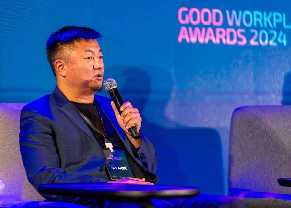 Монголын хөдөлмөрийн зах зээлийн чуулга уулзалт 2024 тайзан дээр сууж байхдаа костюм өмссөн эр микрофонд ярьж байна. Түүний ард дэлгэцэнд "Сайн ажлын байрны шагнал 2024" харагдаж байна. Чанга яригч карттай оосор зүүсэн тэрээр ажлын байранд тулгарч буй бэрхшээлийг шийдвэрлэх шийдлүүдийг санал болгоход гэрэлтдэг. - Business.MN