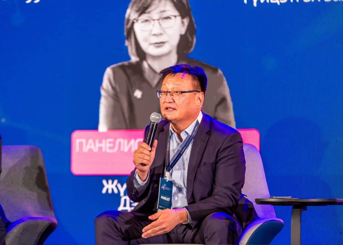 2024 оны Монголын хөдөлмөрийн зах зээлийн чуулга уулзалтын тайзан дээр сууж байхдаа костюм өмссөн эр микрофоноор ярьж байна. Түүний ард эмэгтэй хүний хар цагаан дүрс, зарим текстийг харуулсан том дэлгэц байдаг. - Business.MN